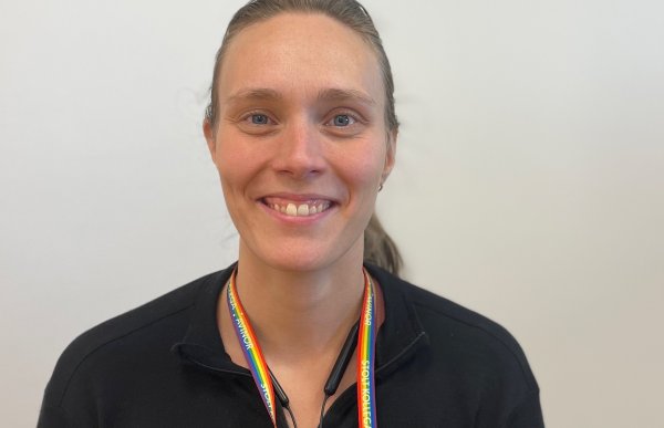 Portrettbilde av en dame med hestehale, svart genser og flerfarget nøkkelring rundt halsen. Hun smiler. I bakgrunnen er en hvit vegg. 