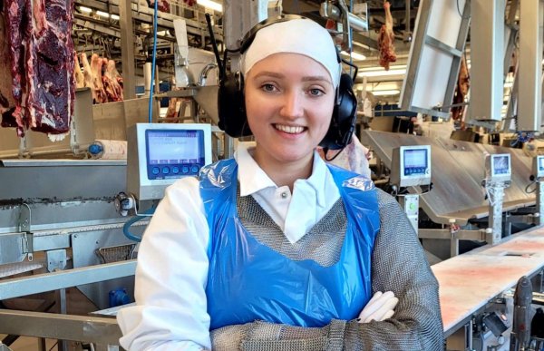 En dame i hvitt arbeidstøy og et blått plastikkforkle. På hodet har hun en hvit lue og hørselsvern. Hun står i et produksjonslokale. Fra taket henger det slakt.