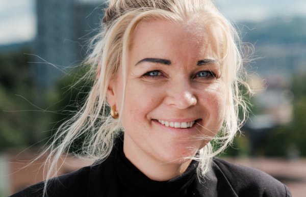 En kvinne med blondt, oppsatt hår og svart genser og jakke. Hun ser rett i kameraet og smiler.