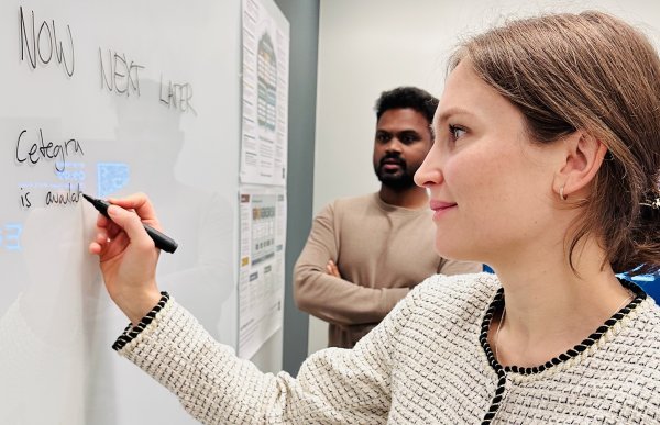Produktleder Kari Clifford på jobb.  En kvinne står ved en whiteboard og skriver med en svart tusj. Hun har håret bundet bak og bærer en lys jakke. Bak henne står en mann og følger med på det hun skriver.
