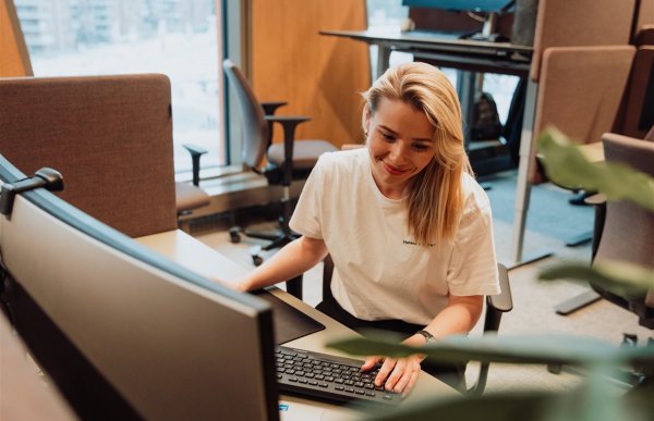 Dame med lyst hår og hvit t-skjorte sitter ved en arbeidspult og jobber på en stor pc