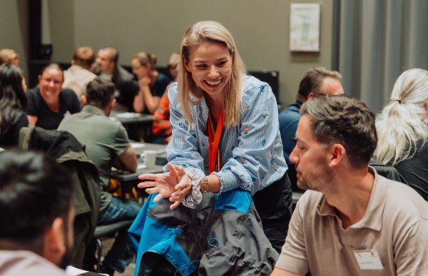 Lyshåret dame med lyseblå skjorte og nøkkelholder rundt halsen står over en stol og prater og smiler til en mann som sitter ved et bord. Det er mange folk i bakgrunnen.