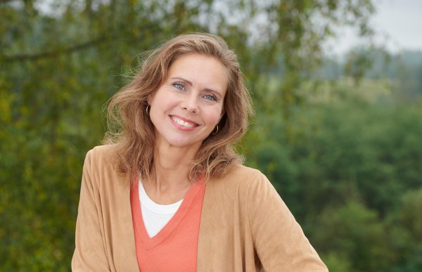 En kvinne kledd i oransje står utendørs med grønn natur som bakgrunn. Hun har lysebrunt, halvlangt bølgete hår og smiler til kameraet.