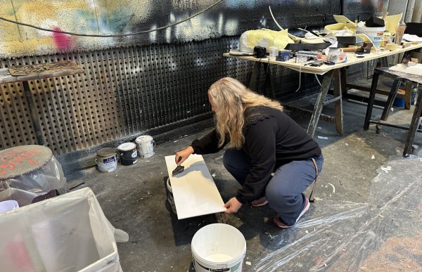 En person med langt, lyst hår sitter på huk i et verksted og påfører hvit maling på en treplate. Rundt ligger malingsspann, og bak er en rotete arbeidsbenk full av verktøy og materialer. Veggen er tilsølt med mange lag maling som gir et kreativt preg.