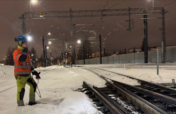En mann med gult og oransje arbeidstøy med refleks, og blå vernehjelm og lykt, står og snakker i telefonen på venstre side av togspor. Det er også elektrisk anlegg over togsporene. Det er vinter og snø. 