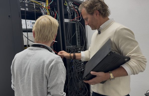 To menn, begge med kort, blondt hår og lyse gensere, står foran et åpent skap med nettverksutstyr og mange kabler. Den ene holder en bærbar PC under armen og peker på en kabel, mens den andre står med ryggen til og ser inn i skapet.