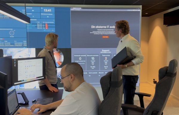 Tre menn befinner seg i et kontorlokale med flere dataskjermer. To av mennene står og ser på en stor skjermvegg med ulike systemoversikter, mens den tredje sitter ved en arbeidsplass foran flere skjermer.