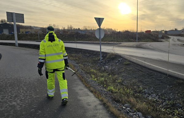 En person i neongult refleksantrekk med logoen «Rejlers» på ryggen går langs en asfaltert vei i lav kveldssol. Personen holder et måleinstrument i hånden. Rundt ham ser vi veikryss, skilt, lysmaster og et åpent landskap med lave bygninger og åser i bakgrunnen. Solen står lavt på himmelen og gir et gyllent lys over området.