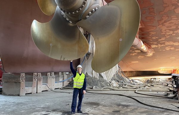 Mann med mørke klær, refleksvest og vernehjelm står under en veldig stor motor i stål