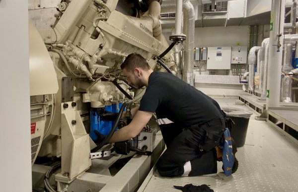 En person arbeider med en stor motor i et teknisk rom. Maskindeler, rør og kontrollskap omgir området der arbeidet utføres.