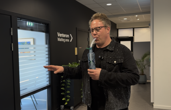 Mann med kort, blondt hår, iført en svart jeansjakke, holder en vannflaske som er halvfull med vann. Et rør stikker opp fra flasken, og han blåser gjennom røret for å varme opp stemmen og løsne slim i halsen før sang.