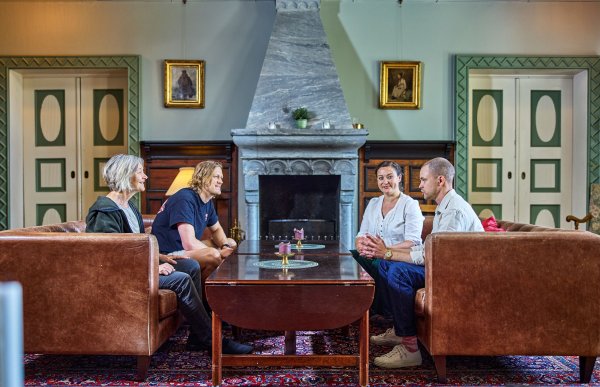 Fire personer sitter i et staselig rom i et hus. Det er en stor peis og fine, grønne dører på hver side. Det er to damer og to menn, de sitter to og to i brune skinnsofaer og prater. 