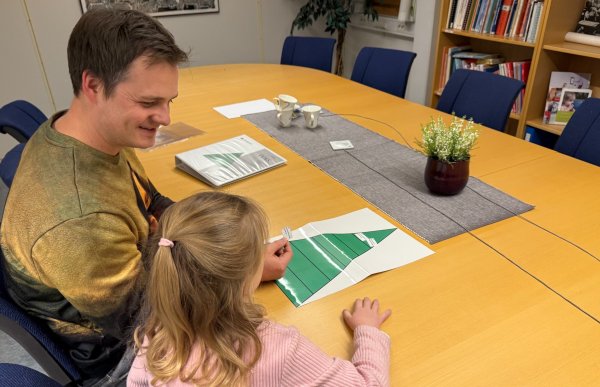 En mann og et lite barn sitter ved et stort møtebord. De ser sammen på et ark med grønne felt som ligger på bordet. Rommet har bokhyller, stoler langs veggen og kaffekopper på bordet, og fremstår som et møterom eller samtalerom.