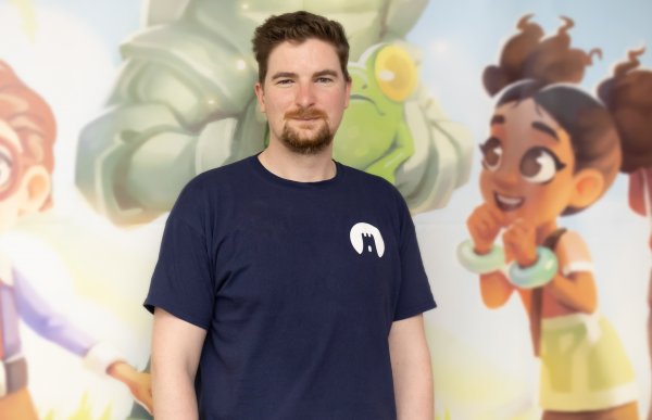 En mann i mørkeblå t-skjorte og beige bukser. Han står foran et stort bilde. Han er i en bygning.