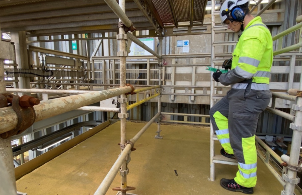 En mann står sidelengs i gule og svarte arbeidsklær med refleks. Han har også på seg hjelm. Han står på et stillas med den ene foten på et trappetrinn. Han holder et skjema og en penn i hånden mens han ser ned.