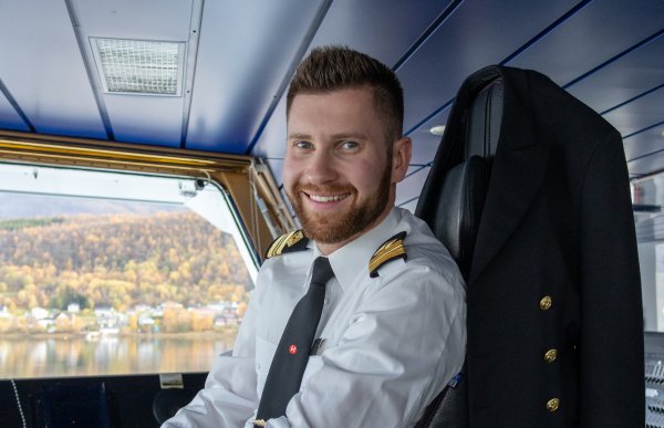 En mann i offiseruniform. Han sitter i en stol på brua på en båt. Han smiler til kamera.