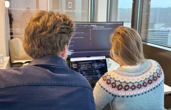 skjerm med programmeringskode. En bærbar PC står foran dem med utviklingsverktøy åpent. De jobber tett sammen i et kontormiljø med glassvegger og utsikt over byen.