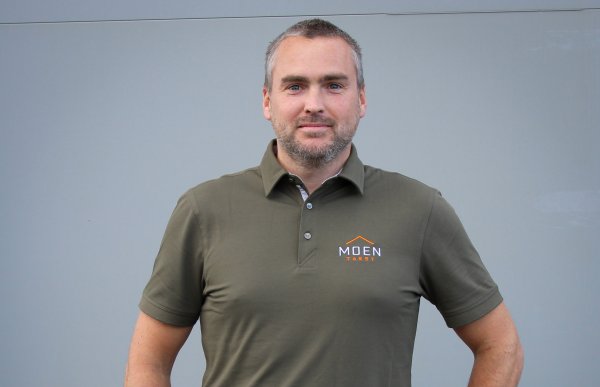 En mann står og ser rett inn i kameraet med et lite smil. Han har hendene på hoftene, og har på seg en kortermet skjorte. Mannen har mørkt og grått kort hår og litt skjegg. 