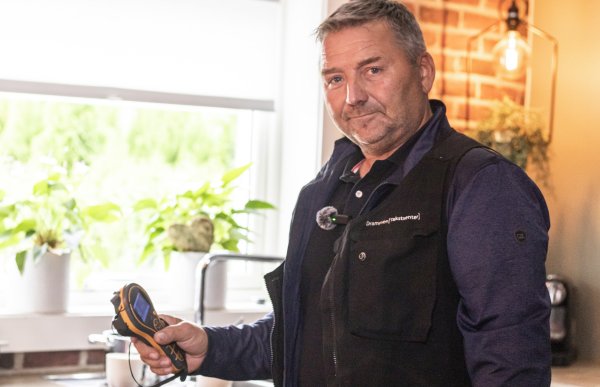 Mann i mørk vest og blå skjorte holder et håndholdt måleinstrument ved en kjøkkenbenk med planter i vinduskarmen bak.