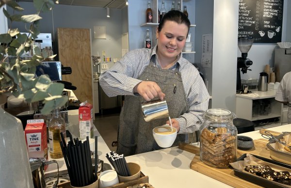 En jente med brunt hår, grått forkle og stripete blå skjorte heller melk i en kaffekopp.. 