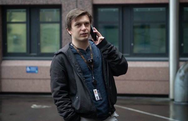 En person med ID-bånd står utendørs foran en bygning og holder en mobiltelefon mot øret, som om de er i en samtale. Personen har på seg jakke og ser litt til siden.
