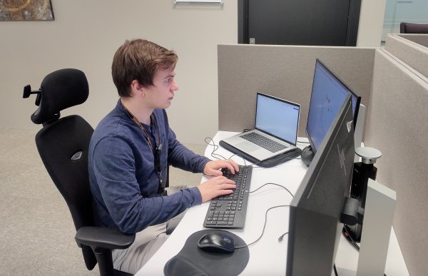 En person sitter ved en kontorpult i et åpent kontorlandskap og jobber på en datamaskin. Han ser på skjermen mens han skriver på et tastatur, med en laptop og en ekstra skjerm ved siden av.