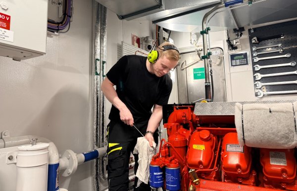 En mann i arbeidsklær og hørselsvern står bøyd over en stor rød motor i maskinrommet på en båt. Han holder en klut og arbeider med vedlikehold på motoren. Rundt ham er det verktøy, rør og teknisk utstyr montert på vegger og i tak.