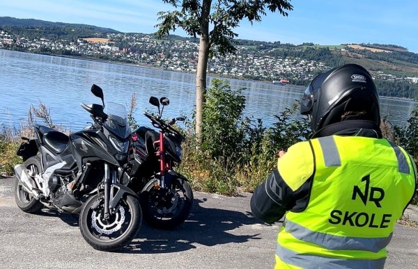 En person med svart hjelm og gul refleksvest med teksten «NR Skole» sitter på en motorsykkel og ser utover en innsjø. To motorsykler står parkert ved siden av hverandre på en utsiktsplass. På andre siden av vannet ligger en by med hus og bygninger.