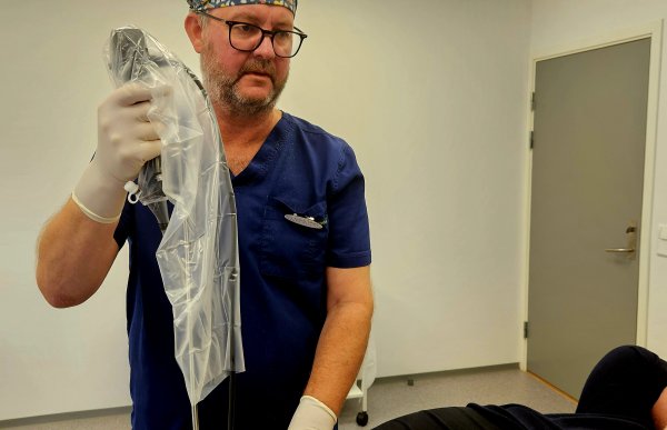 En mann i blå legeoverdel med en mønstret lue og briller. Han holder et apparat over en person som er delvis skjult.