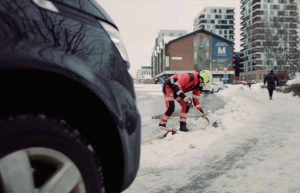 En person med arbeidsklær står og graver i snøen ved siden av en bilvei. 