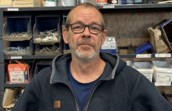 En mann med kort grått hår, skjegg og briller står foran hyller med bokser, sprayflasker og ulike deler. Han har på seg en mørk blå fleecejakke over en blå T-skjorte og ser mot kameraet.