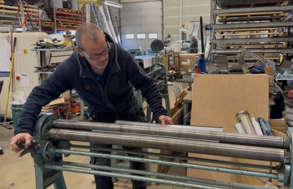 En mann med briller og mørke arbeidsklær arbeider ved en stor valsemaskin i et verksted. Han holder i et håndtak på maskinen og fører en metallplate mellom de lange valsene. Rundt ham står maskiner, hyller og materialer.