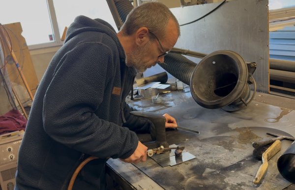 En mann med briller og mørk blå fleecejakke bøyer seg over en arbeidsbenk i et verksted. Han bruker en gassbrenner på en liten metallplate som ligger på benken. Rundt ham ligger ulike verktøy og deler i metall.