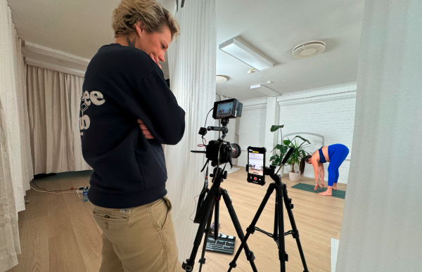 Et rom med flere hvite laken hengende rundt. En person, videografen, står og følger med på bilde på kameraet i som står plassert på et stativ i rommet. . En annen person i tettsittende blått og sort treningstøy står fremoverlent på gulvet i rommet.