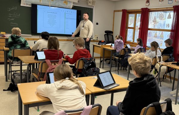 En mann står foran en digital tavle i et klasserom. Det sitter barn på pultene med iPad på pulten. En elev rekker opp hånden.