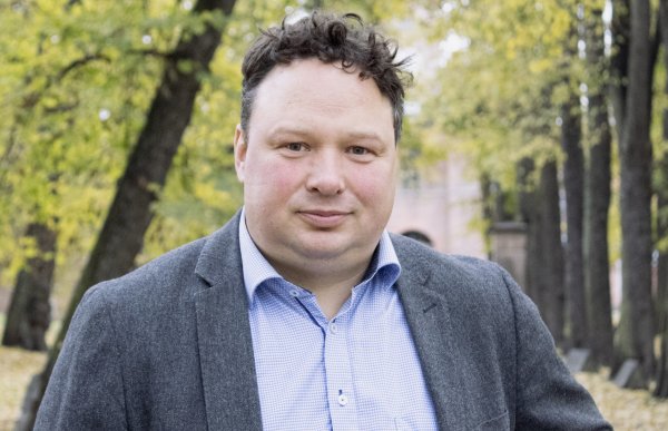 En mann i blå skjorte og grå jakke ser rett i kameraet. Han er utendørs, og i bakgrunnen er det trær.