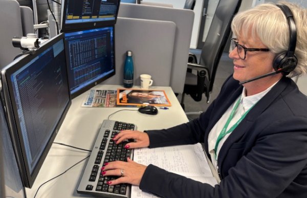 Dame med lys skjorte og svart blazer sitter med et headset på hodet og skriver på en pc. Det står to pc-skjermer foran henne.