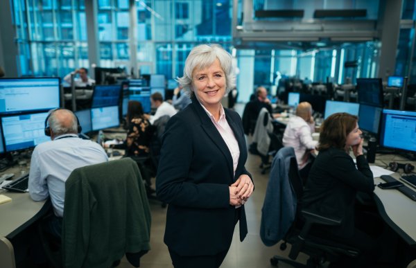 Dame med lys skjorte og svart blazer står midt blant mange mennesker på et stort kontorfellesskap og smiler til kamera. Folk rundt følger med på pc-skjermene.
