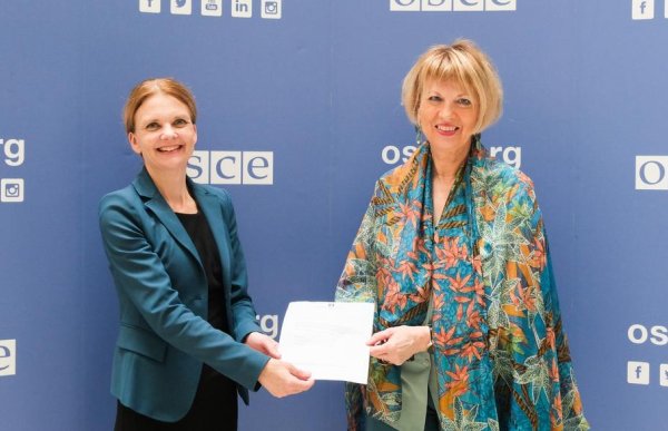 To kvinner står side om side foran en blå bakgrunn med gjentatte logoer og teksten «OSCE». Begge smiler mot kamera. Kvinnen til venstre har på seg en mørk kjole med en grønn blazer, og holder et dokument som hun rekker over til kvinnen ved siden av. Kvinnen til høyre har kort, lyst hår og er kledd i en fargerik, mønstret sjal eller jakke over lyse klær. De holder dokumentet sammen, som om det overrekkes i en formell sammenheng.