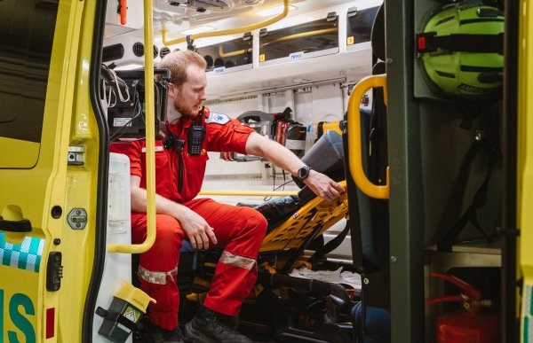 En mann i rød uniform synes gjennom sidedøren på en ambulanse. Han sitter i et sete i bakrommet, og tester ryggstøtten på båren.