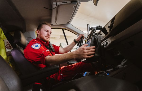 En mann i rød uniform sitter i førersetet på en ambulanse. Vi ser ham nedenfra, og han ser på en touchpad på midten av dashbordet.