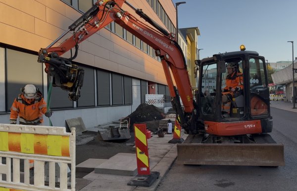 To personer i oransje arbeidsklær og hjelm arbeider på et anleggsområde ved en bygning. En av dem, en kvinne, sitter i førerhuset på en oransje gravemaskin, mens den andre personen står ved siden av og holder en grønn løftestropp som henger fra gravemaskinen.