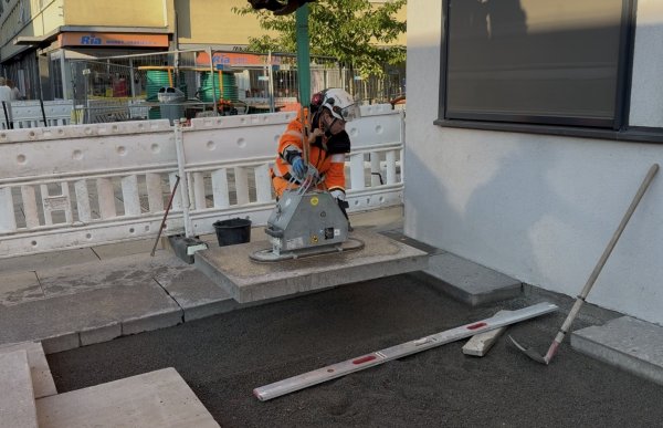 En kvinne i oransje arbeidsklær, hjelm og hansker står ved en stor steinhelle som henger i grønne løftestropper. Hun holder i stroppene og styrer hellen på plass på bakken. Rundt arbeidsområdet står hvite sperregjerder, og et vater og annet arbeidsutstyr ligger på bakken.