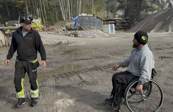 To menn befinner seg på et gruslagt anleggsområde. Mannen til venstre står i svart arbeidstøy med gule refleksfelt på buksen. Mannen til høyre sitter i rullestol og har på seg grå genser, svart bukse, lue og hørselsvern. Rundt dem ligger hauger med stein og grus, og i bakgrunnen ses blant annet en container og en fjellvegg.