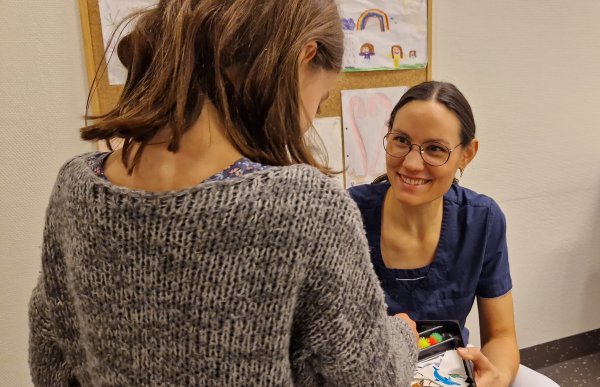 En dame med mørkt hår og briller, ikledd blå t-skjorte smiler til en ung jente med grå genser og brunt hår. Det er tegninger på veggen i bakgrunnen.