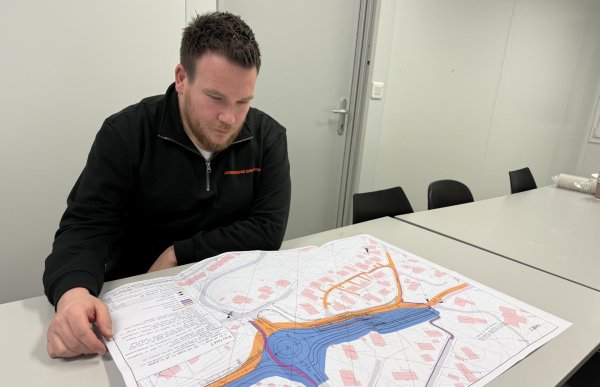 En mann med brunt hår og skjegg sitter ved et bord og studerer et stort kart. Han har på seg mørkt arbeidstøy. Bildet er tatt innendørs i et møterom.