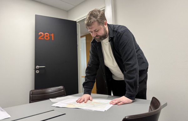 En mann med brunt hår og skjegg står lent over et møtebord og studerer flere store ark som ligger utover bordflaten. Han har på seg mørk jakke og lys t-skjorte. I bakgrunnen sees et kontorrom med stoler og en svart dør merket med tallet 281.