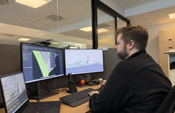 En mann med brunt hår og skjegg sitter ved en arbeidsplass med flere skjermer og jobber på datamaskin. Skjermene viser digitale kart og illustrasjoner, og på pulten står tastatur, laptop og kontorutstyr. Kontoret har glassvegger og gir inntrykk av et moderne arbeidsmiljø.