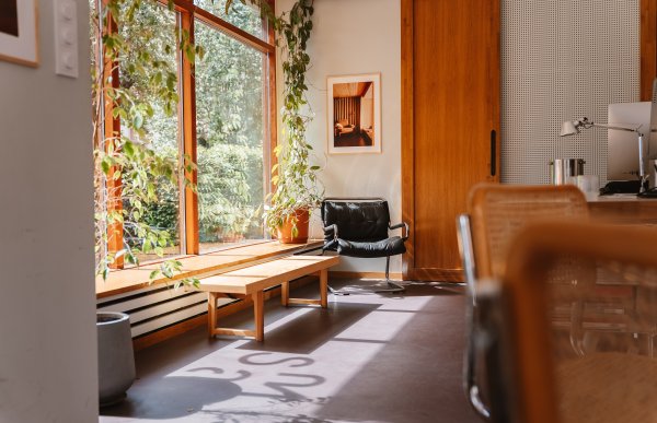 Et illustrasjonsbilde fra arkitektkontoret viser en benk i tre, og en liten svart skinnstol, som står ved karmen av et stort vindu. Det er grønne planter rundt vinduet, og solen skinner inn i rommet.