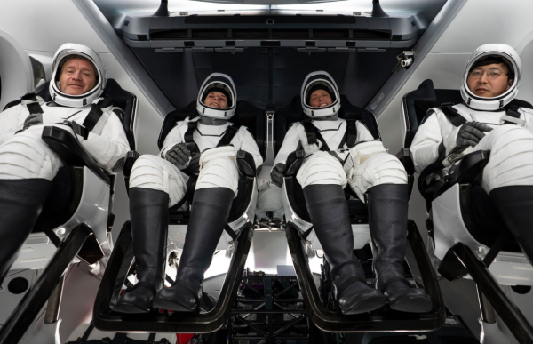 Fire astronauter sitter side om side inne i en romkapsel. De har på seg hvite romdrakter med svarte detaljer og hjelm. Alle sitter bakoverlent i mørke seter med beina støttet opp på fotstøtter. Interiøret i kapselen er lyst og moderne.